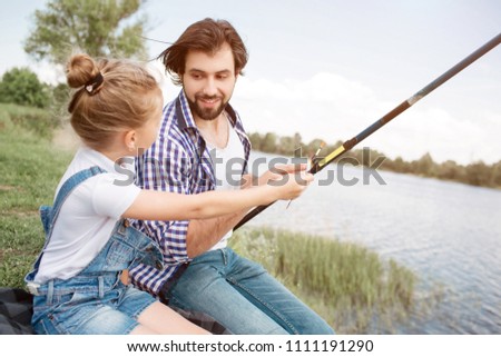 A picture of girl asking his dad about fishing for a bit. She wants to take fish-rod into her hands and do it by herself. Guy is looking at her and giving her fish-rod. He loves his daughter.