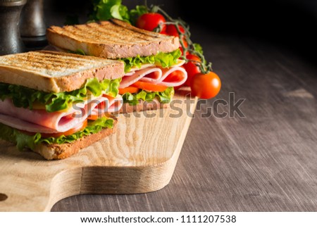 Close-up of two sandwiches with bacon, salami, prosciutto and fresh vegetables on rustic wooden cutting board. Club sandwich concept.