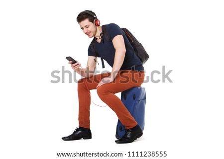 Portrait of young man sitting on suitcase and looking at mobile phone while listening to music 