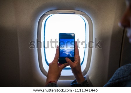 Young woman in plane taking photo of clouds and sky as seen through window on her smartphone. Focus on phone. Travel and technology