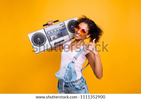 Yes, done! Portrait of trendy stylish girl in eyewear holding boom box on shoulder gesturing thumb up sign looking at camera isolated on yellow background. Recommend choice advice concept Royalty-Free Stock Photo #1111352900