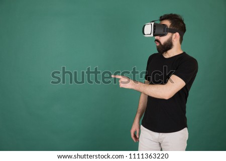 Young man wearing a casual outfit and wearing a VR glasses and looking so shocked and pointing by his fingers, standing on a green background