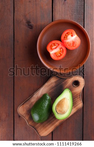 Avocado on an old wooden board, halved avocado and tomato on a wooden background, green tropical fruit top view, red and green vegetables on a vintage background, copy space