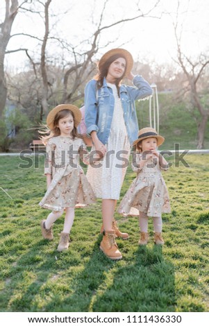 Young stylish pretty woman dressed summer dress, denim jacket and hat walking with her two little charming daughters in similar summer outfits in sunny summer park. Happy family portrait outside
