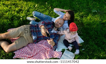 Happy family is laying on the pled and doing selfie with a baby at sunset in the park. Father and mother take pictures of themselves with the baby, camera movement