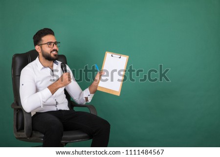 Young interviewer sitting on the chair holds a clipboard showing it talking on the microphone smiling on a green background.