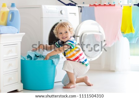Child in laundry room with washing machine or tumble dryer. Kid helping with family chores. Modern household devices and washing detergent in white sunny home. Clean washed clothes on drying rack.  Royalty-Free Stock Photo #1111504310