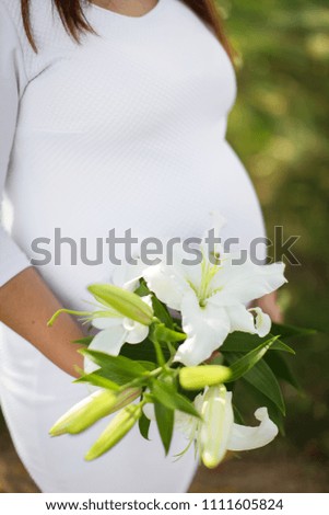 close up pregnant girl in white dress holds a bouquet of white lilies next to a pregnant tummy.