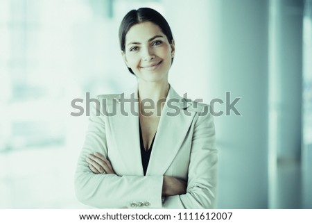 Closeup portrait of smiling beautiful middle-aged business woman wearing jacket and standing in light office hall with her arms crossed. Front view.