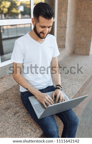 Portrait of a beautiful stylish guy dressed in a white t-shirt, who sits on a parapet, works with a laptop, on the street against a glass building,