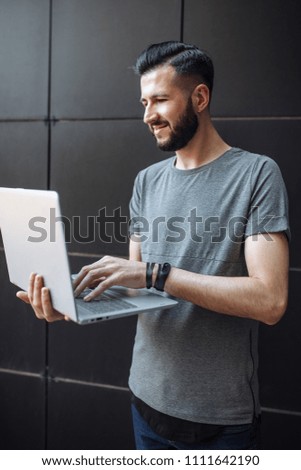Portrait of handsome stylish guy, the hipster, working on laptop, wearing a gray blank t-shirt standing against a black wall.