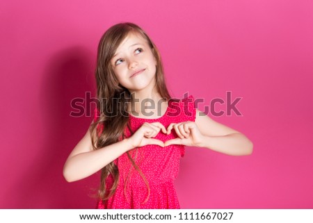 Little 8 years old girl make heart gesture with her hands on a pink neutral background. She has long brunette hair and wear red summer dress. Funny expression on her face