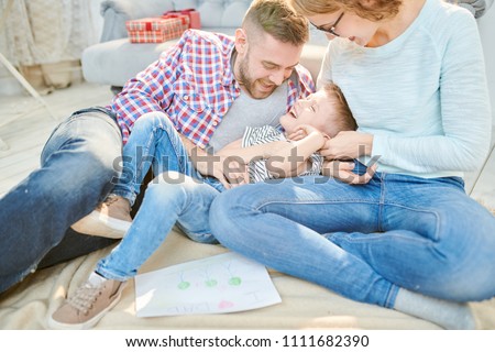 Adorable family of three enjoying each others company while gathered together at cozy living room illuminated with sunbeams, loving dad tickling his cute little son