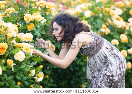 Young Caucasian women with dark curly hair taking picture of yellow rose in a rose garden with a black smartphone 