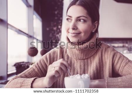 Dreamful young woman in sweater drinking hot chocolate in cafe. Smiling woman looking aside when she sitting at table. Beautiful woman stirring sugar into cup. She waiting for friend. Leisure concept