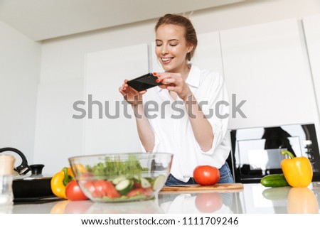Smiling young woman taking picture with mobile phone while cooking salad on a kitchen