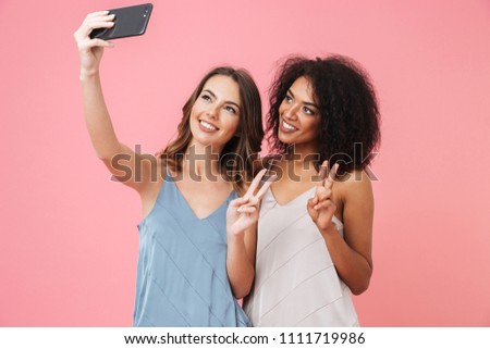 Two pretty young girl in dresses taking a selfie while showing peace geture isolated over pink background