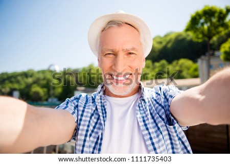 Self portrait of cheerful positive man in straw hat with beaming toothy smile shooting selfie on front camera over blurred street background