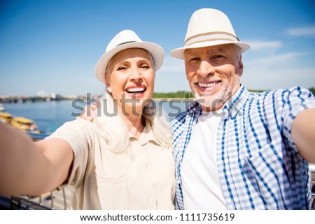 Portrait of stylish trendy senior couple in casual outfits straw hats shooting selfie on front camera over blurred river background having good mood enjoying sunny day sunshine