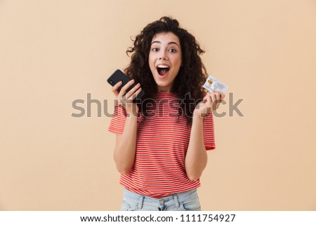 Photo of emotional excited pretty young curly woman isolated over beige background using mobile phone holding credit card.