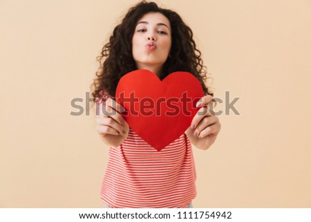 Photo of happy young woman isolated over beige wall background looking camera holding heart in hands blowing kisses.