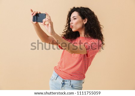 Portrait of a beautiful young girl with curly hair taking selfie with mobile phone isolated over beige background