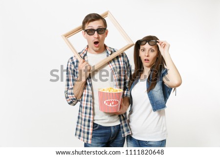 Young shocked couple, woman and man in 3d glasses and casual clothes watching movie film on date, holding bucket of popcorn and picture frame isolated on white background. Emotions in cinema concept
