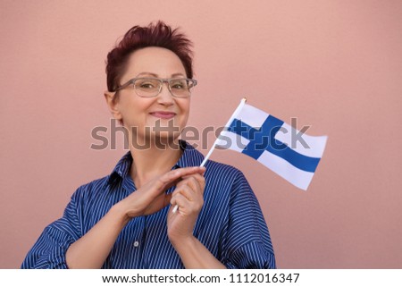 Portrait of middle aged woman holding flag of Finland.  Patriotic lady 40 50 years old with national Finnish flag over pink wall background.  Learning/studying Finnish language. Visit Finland concept