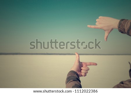 Hands of a man in camouflage showing a square against the background of the icy lake with horizon.