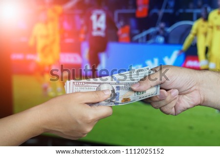 Football Gambling Concept : Hand gives money to another over TV screen showing football match as background.