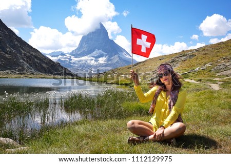 Happiness Asian beauty young tourists sitting and smiling with hand holding Swiss flag near the alpine lake of Riffelhorn in front of mountain Matterhorn peak, Zermatt, Switzerland.Summer Vacation  Royalty-Free Stock Photo #1112129975