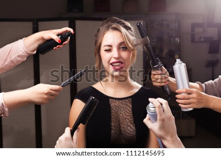 beautiful emotional woman is surrounded by many hands holding hair curler, sprays and brushes. girl is showing a tongue with one eye closed. concept of professional makeup products Royalty-Free Stock Photo #1112145695