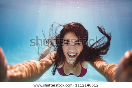 Woman swimming underwater in pool smiling and making a selfie. Young female swimmer shooting herself while swimming underwater.