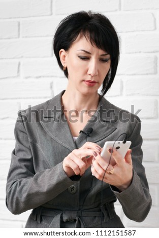 business woman dressed in a gray suit dialing on smartphone or preparing to online broadcasting, stay in front of a white wall