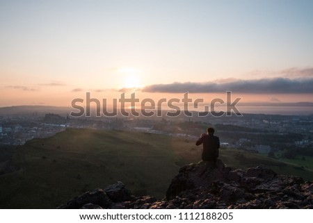 A young man taking a picture and enjoyng the view of Edinburgh from the top of Arthur's Seat in the middle of Holyrood Park