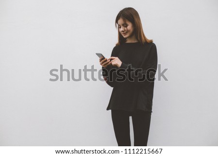 Photo of a smiling happy hipster girl wearing a black blank hoodie using a mobile smartphone while standing on a white concrete wall background. Young woman typing via cellphone