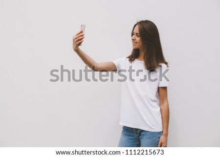 Smiling happy hipster girl wearing a casual white t-shirt and taking selfie while standing on a white concrete wall background