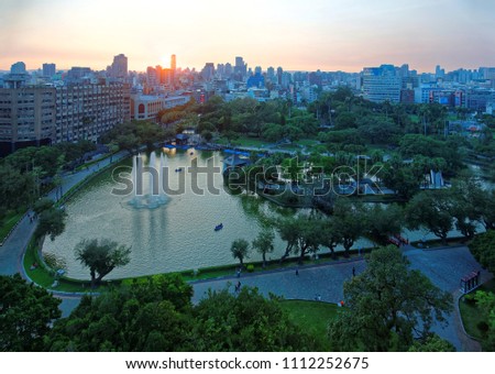 Sunset scenery of Downtown Taichung, a vibrant city in central Taiwan, with the golden sun setting behind modern skyscrapers and a pond surrounded by green forests in an urban park under twilight sky