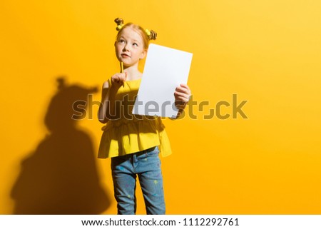 Girl with red hair on a yellow background. A beautiful girl is holding a pencil and a white sheet in her hands. The child pondered.