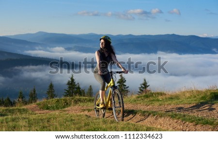Young female cyclist riding on yellow bicycle on a rural trail in the mountains, wearing helmet, enjoying sunny morning. Foggy mountains, forests on the blurred background. Outdoor sport activity
