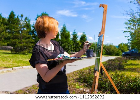 A young blonde female artist in a black T-shirt and jeans paints on canvas and an easel with oil paints in a city park on a sunny day. Drawing on an open air