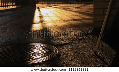 A damp street is mildly illuminated by the streetlight casting shadows on the asphalt and a manhole cover.