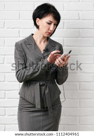 business woman dressed in a gray suit dialing on smartphone or preparing to online broadcasting, stay in front of a white wall