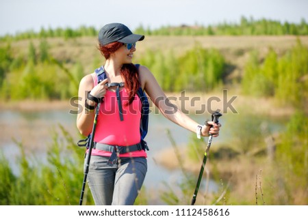 Photo of young sporty girl looking away with walking sticks on background of lake and green vegetation