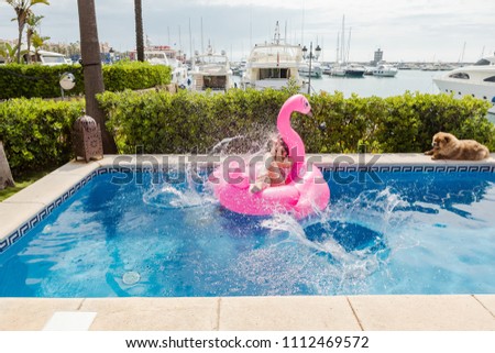 Young woman setting on a inflatable pink flamingo, having sunbathe and closing from water splashes when somebody jumping to the pool 