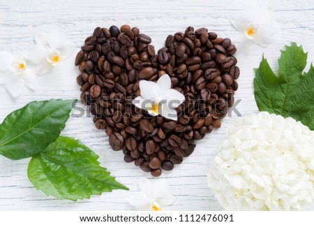 Heart of coffee beans laid out on a white wooden background with white hydrangea flowers and orchids