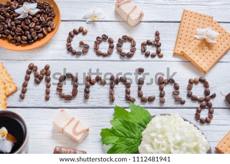 The phrase "Good Morning" is laid out with coffee beans on a wooden background, a cup of coffee, a white hydrangea, orchids and sweets.