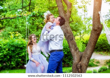 Happy father kisses his little daughter, beautiful wife in blue dress rides on swing, on background of summer park