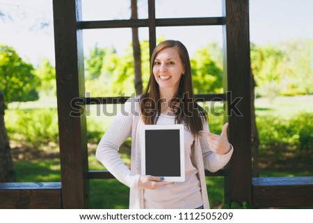 Portrait of beautiful woman wearing light casual clothes. Girl showing camera tablet pc computer with blank empty screen to copy space in park in street outdoors on spring nature. Lifestyle concept