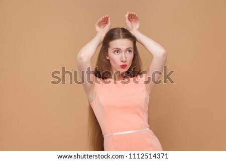 funny girl with bunny style hands. portrait of emotional cute, beautiful woman with makeup and long hair in pink dress. indoor, studio shot, isolated on light brown or beige background.